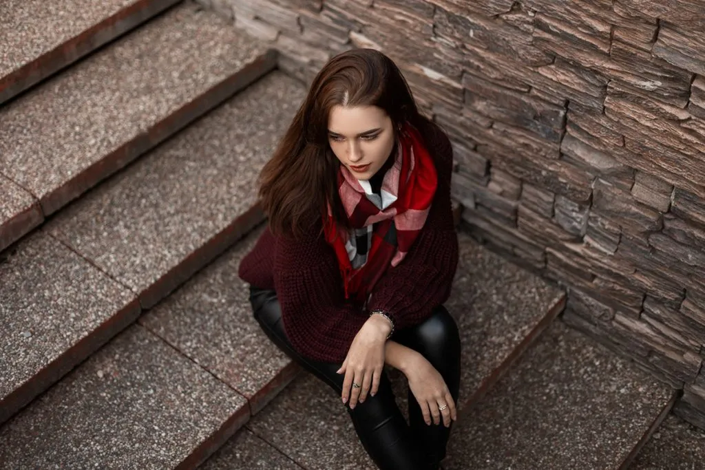 woman in a fashionable knitted vintage sweater in leather pants with a plaid scarf is sitting in the city on a stone staircase