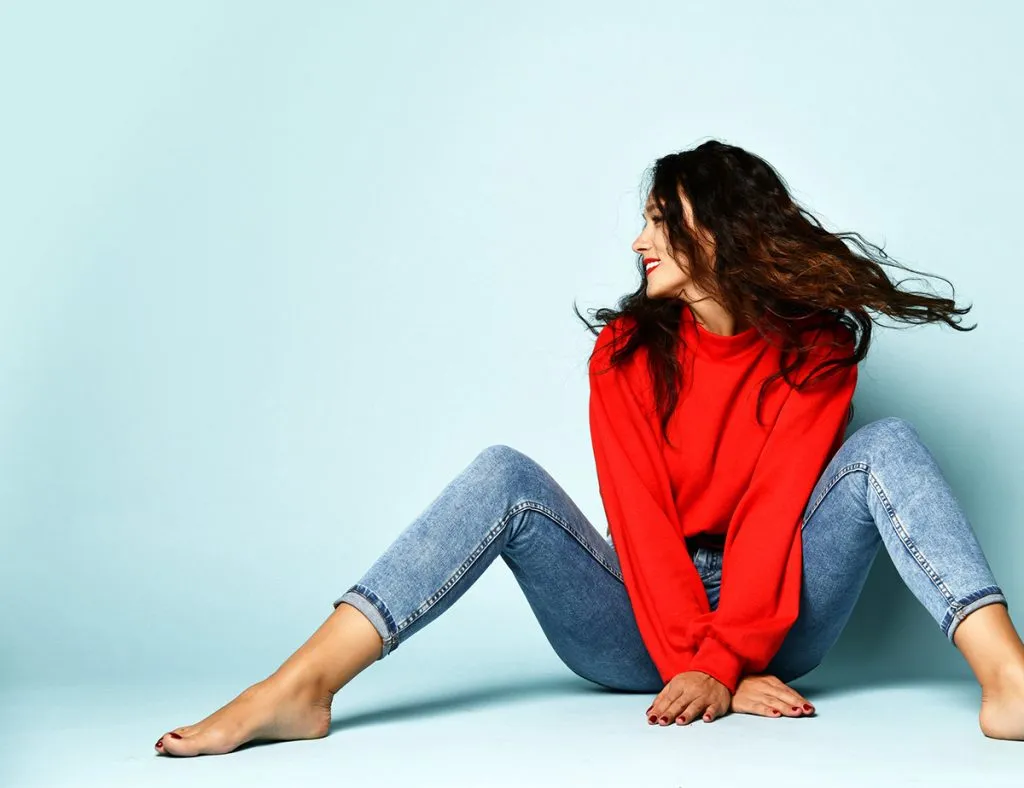 smiling adult woman with long curly brunette hair in red sweater and blue tight jeans is sitting on ther floor looking aside