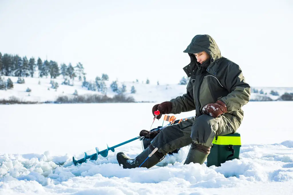 young man wearing a wader is fishing from a hole on ice