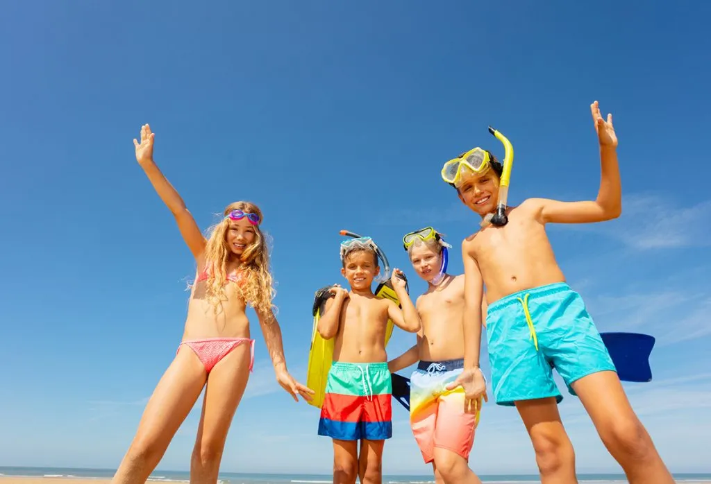 On the beach, a group of happy boys and girls stand together waving their hands in swimwear