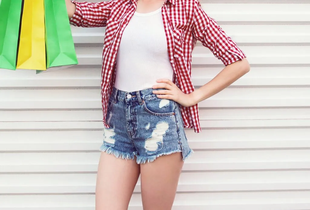 young woman in summer with colorful shopping bags, round straw hat, checkered shirt, and shorts on a white wall background
