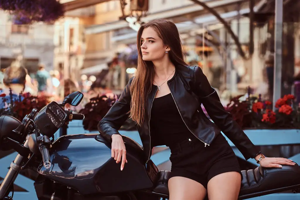 sensual young girl model dressed in a black dress and leather jacket sitting on custom-made motorcycle, posing against terrace of a cafe