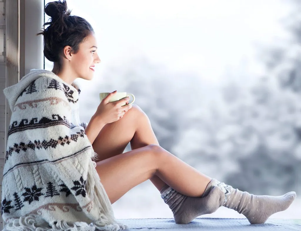 young woman in a knitted Nordic print poncho drinking coffee while sitting at home by the window