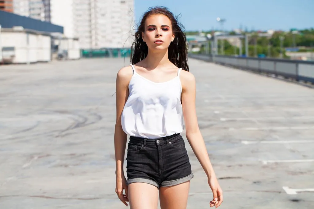brunette model in white summer blouse and denim shorts walking on summer street
