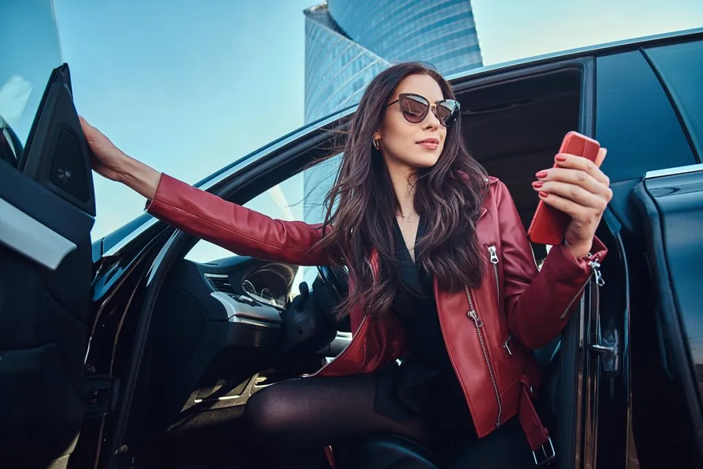 Beautiful smart women is posing in her new car while chatting on mobile phone. She is wearing red leather jacket and sunglasses.