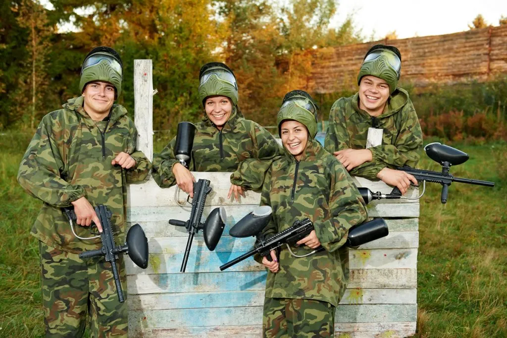 happy paintball sport player girl in protective camouflage uniform and mask with marker gun outdoors