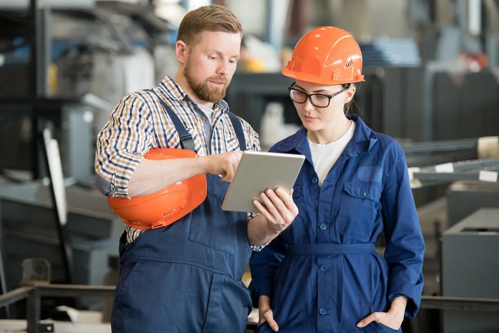 female engineer in uniform and helmet looking at tablet display while listening to colleague explanation of data in warehouse