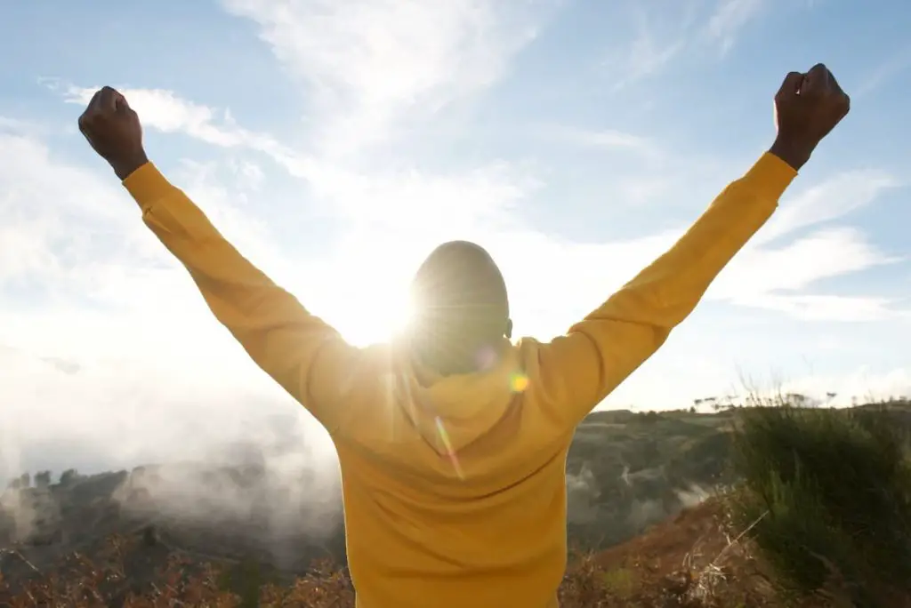 african american man in hoodie with hands raised in air and sun