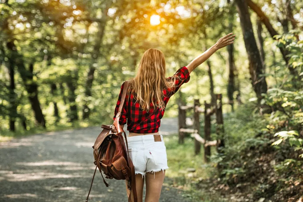 young woman walking on forest road with backpack with arm raised wearing red t-shirt and sexy white shorts