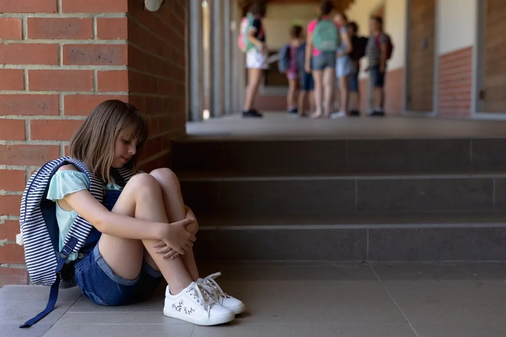 schoolgirl wearing denim shorts, sports shoes and a rucksack sitting on the ground alone in the schoolyard at elementary school looking sad, with other schoolchildren standing together in the background