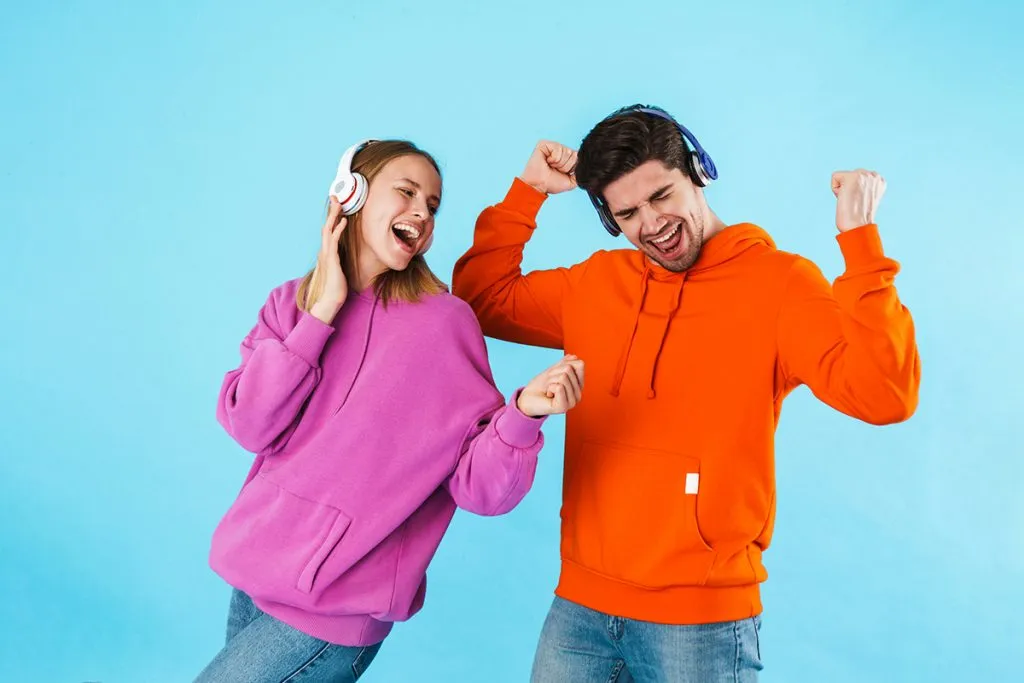 cheerful young couple wearing hoodies standing isolated over blue background, listening to music with wireless headphones, dancing