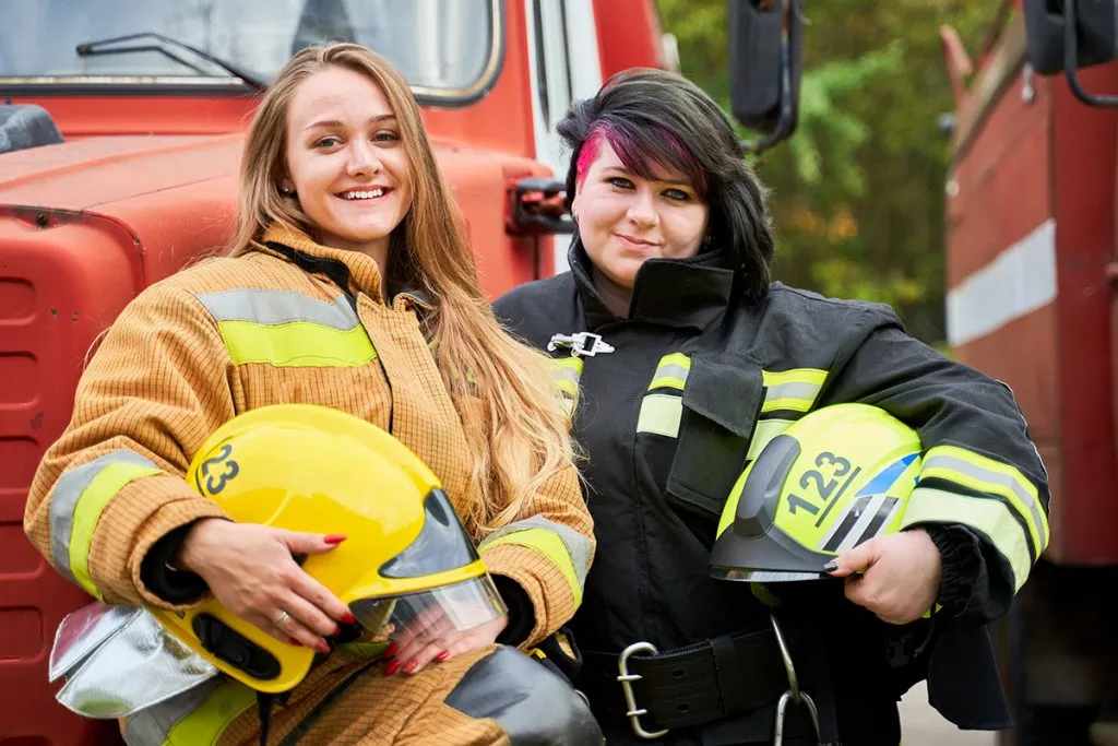 two women firefighter with helmet in his hands standing next to firefighters car