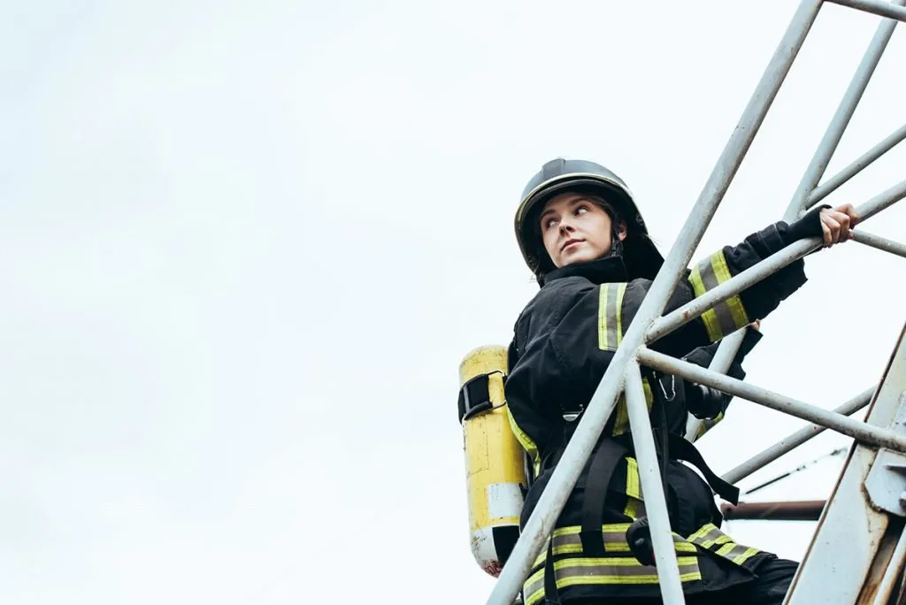 female firefighter in protective uniform and helmet with fire extinguisher on back standing on ladder with blue sky on background