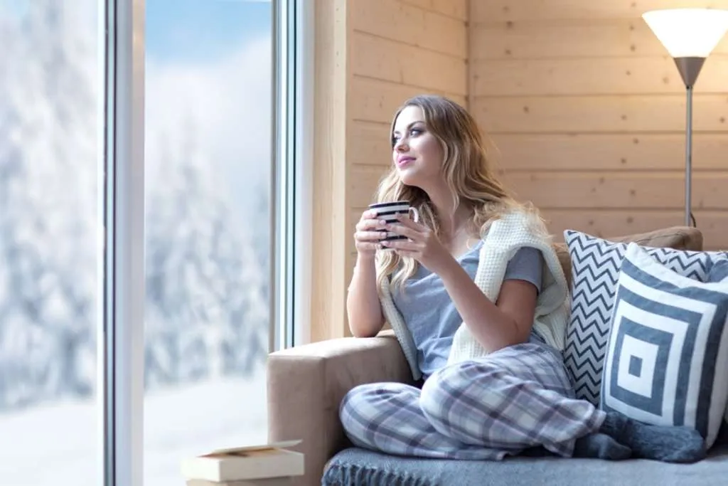 blonde woman with cup of coffee sitting home in living room by the window