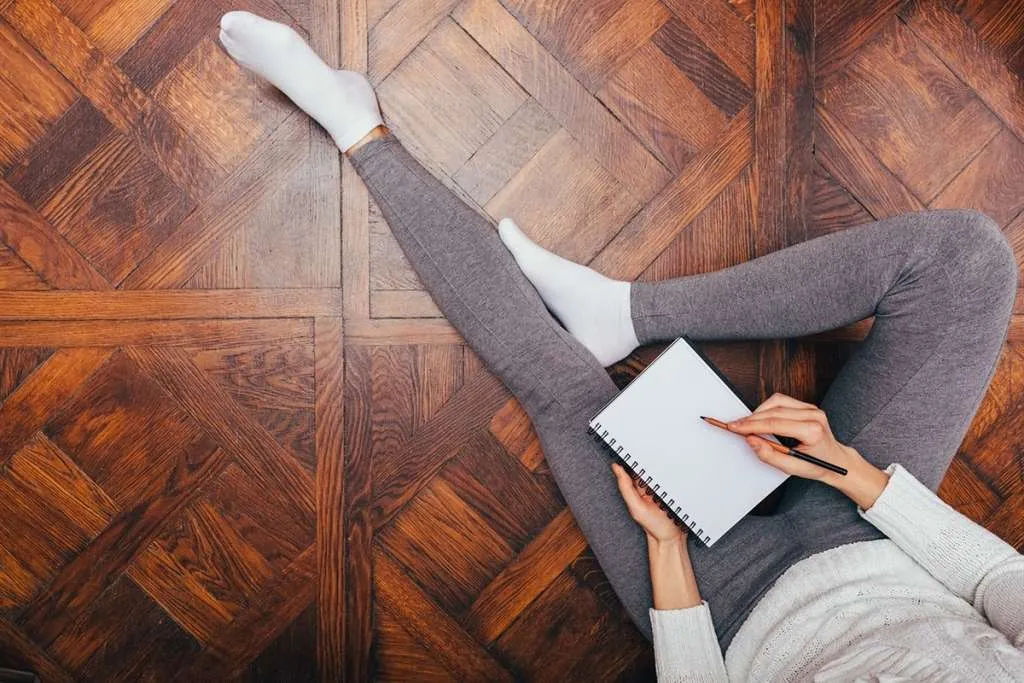 woman in leggings sits on a wooden floor, writing on a notepad